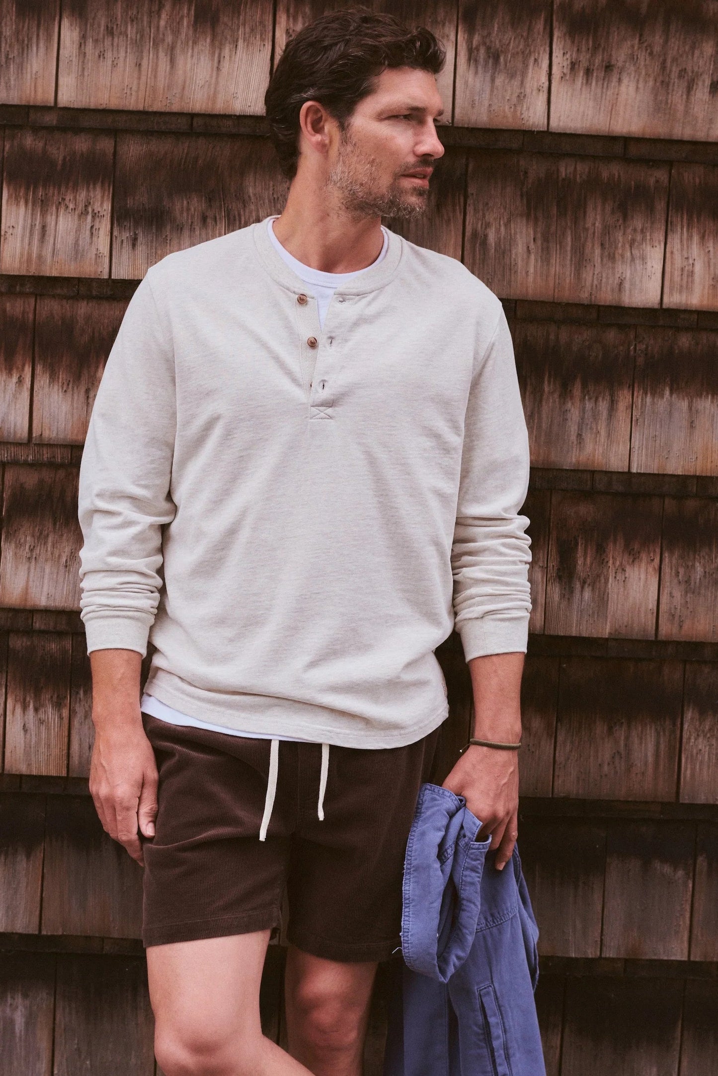 A man wearing a light beige long-sleeve henley shirt and dark brown shorts, standing against a rustic wooden shingle wall and holding a blue shirt.