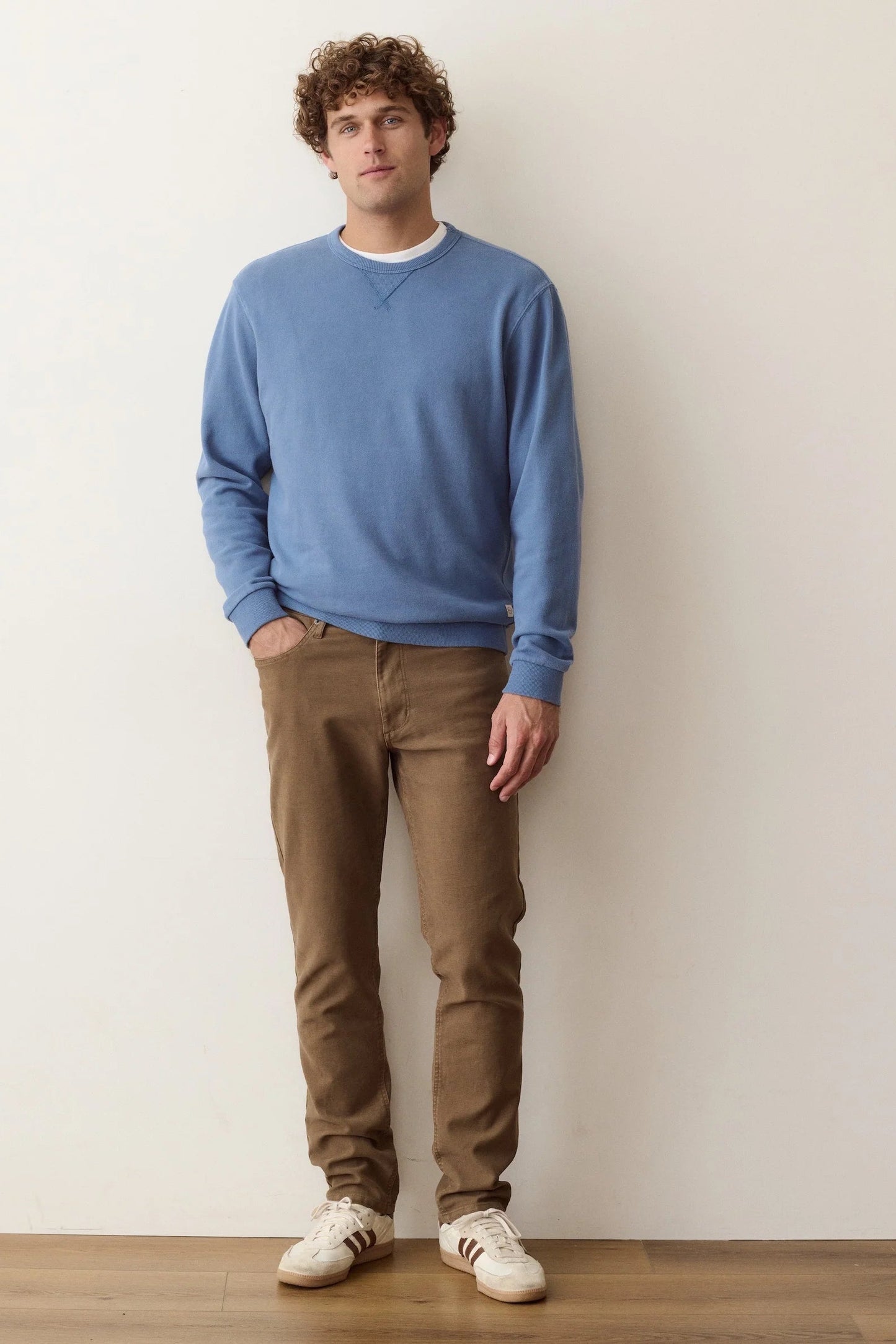 A man stands against a plain off-white wall wearing a dusty blue crewneck sweater, brown trousers, and white sneakers.