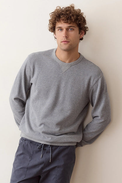 A man with curly brown hair wearing a textured gray crewneck sweatshirt and dark blue drawstring pants, leaning against a plain off-white wall.