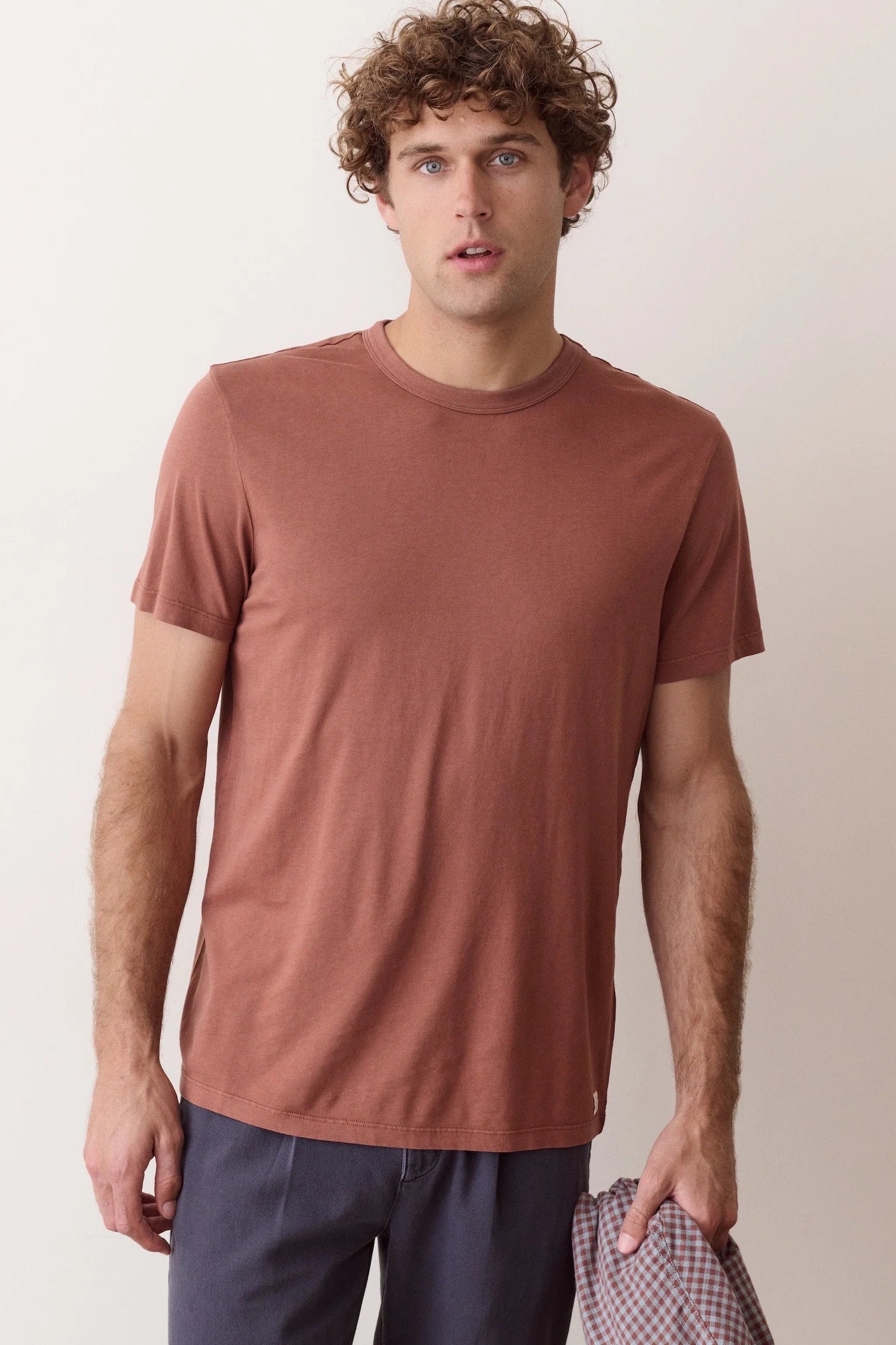 A man with curly brown hair wearing a muted terracotta short-sleeve crewneck t-shirt and navy pants, holding a checkered shirt in one hand against a plain light beige background.