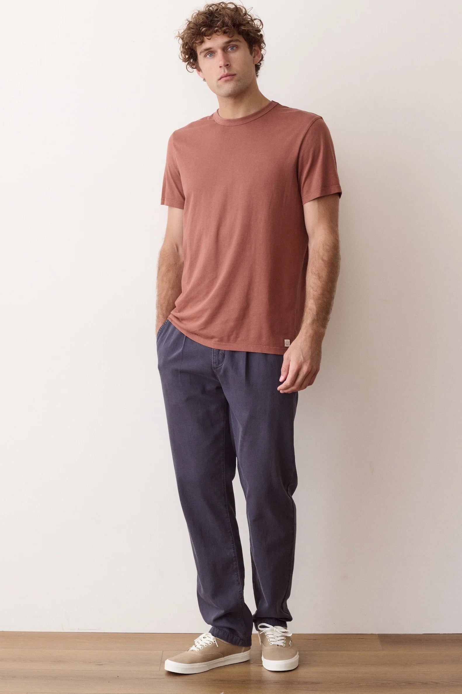 A man wearing a muted terracotta short-sleeve t-shirt, dark navy pants, and beige sneakers, standing against a plain off-white wall.