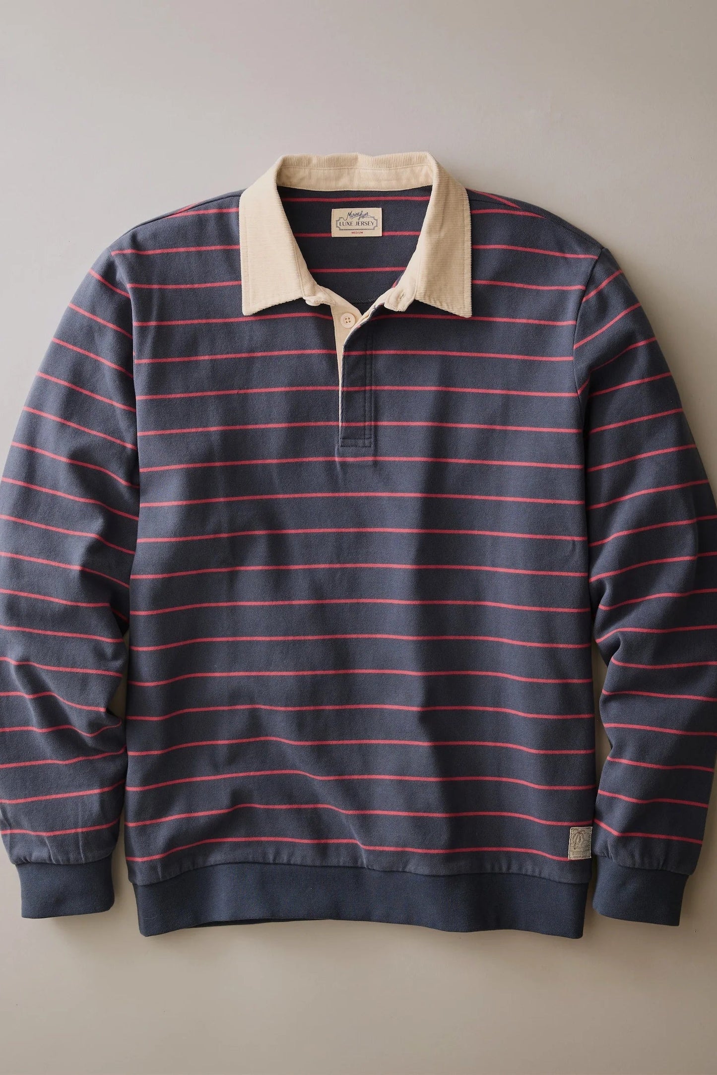 A long-sleeve rugby-style polo shirt with horizontal navy and red stripes, featuring a contrasting beige collar and a partial button placket, displayed flat against a neutral gray background.