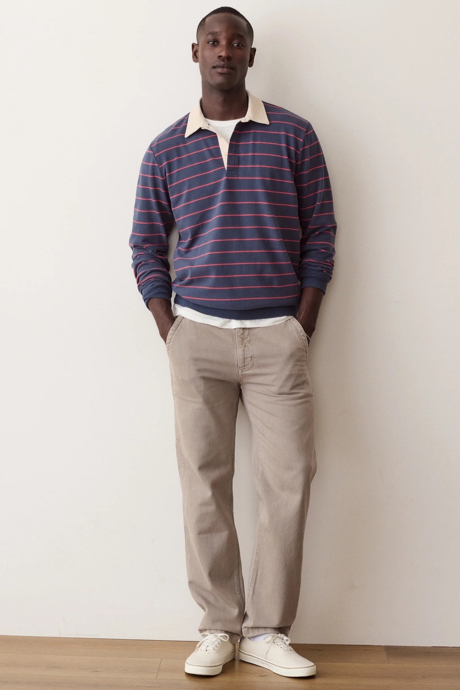 A man stands against a plain off-white wall wearing a navy and red striped long-sleeve polo shirt with a cream collar, beige trousers, and white sneakers.