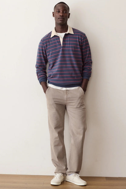 A man stands against a plain off-white wall wearing a navy and red striped long-sleeve polo shirt with a cream collar, beige trousers, and white sneakers.