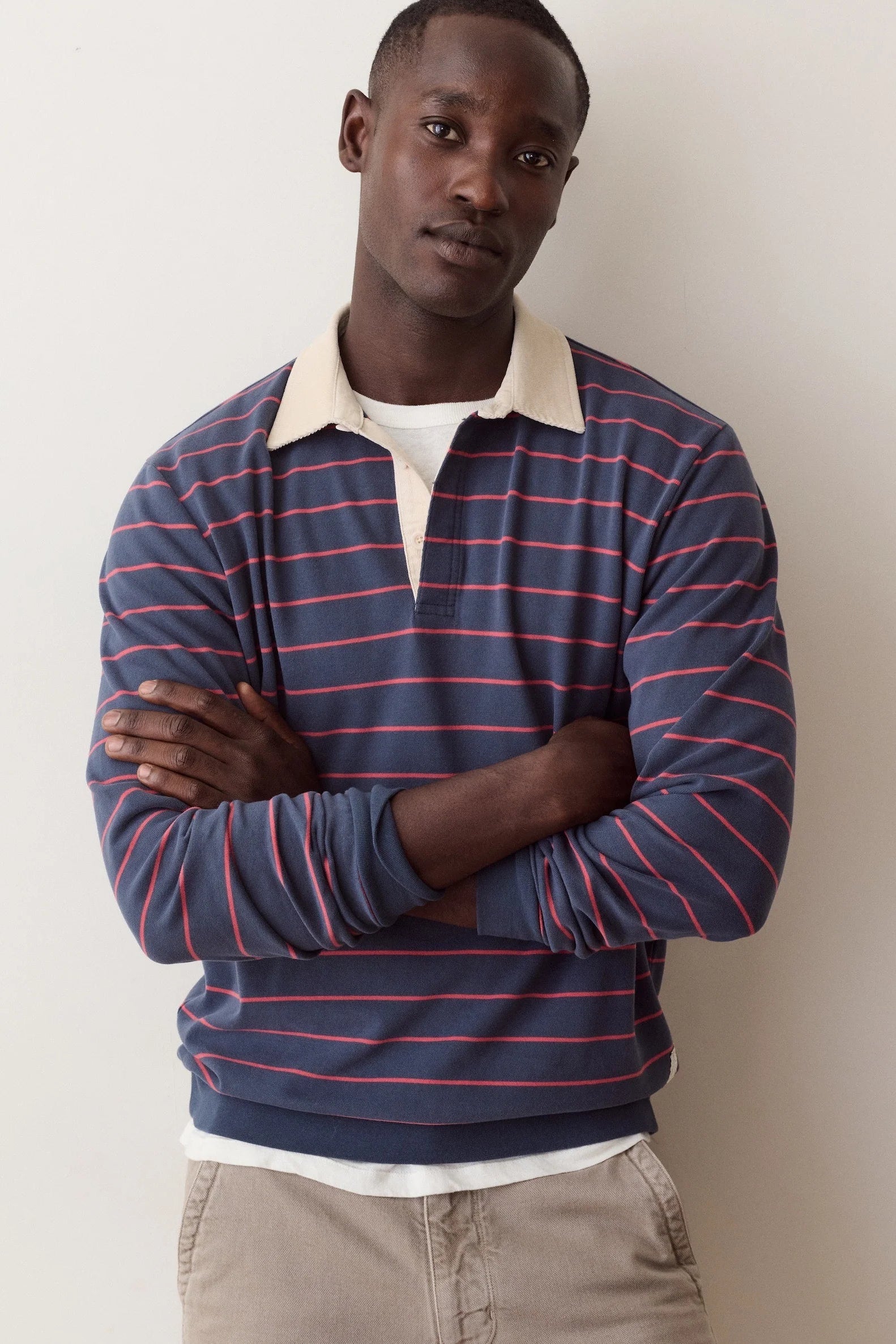A man wearing a navy blue long-sleeve rugby-style sweater with thin red horizontal stripes, a cream collar, and a white undershirt, standing against a plain off-white wall.