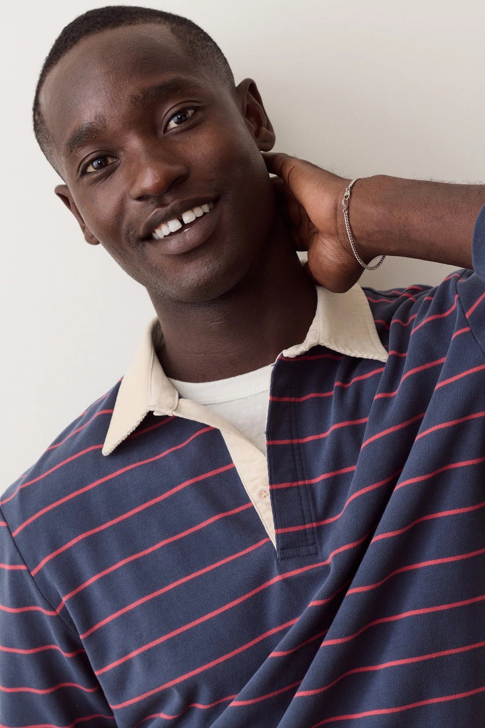 A smiling man wearing a navy blue polo shirt with thin red horizontal stripes, a cream-colored collar, and a white undershirt, posing against a plain off-white background.