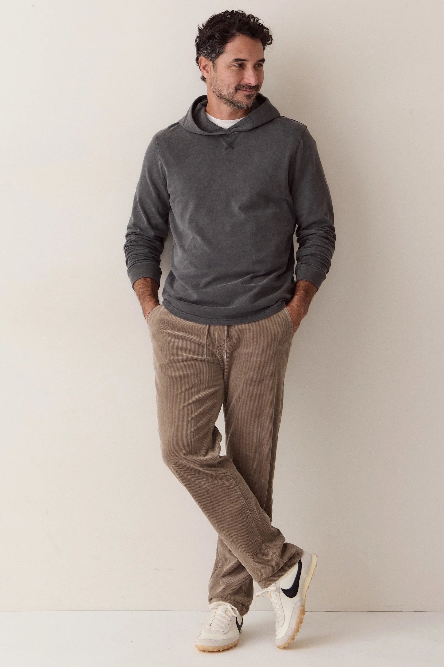 A man wearing a dark gray long-sleeve hoodie, brown corduroy pants, and white sneakers, leaning against a plain off-white wall.