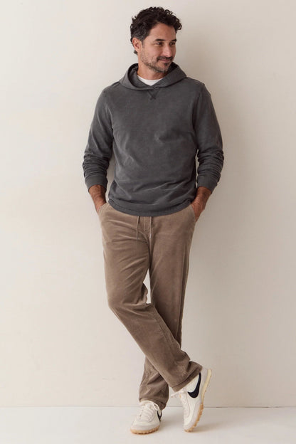 A man wearing a dark gray long-sleeve hoodie, brown corduroy pants, and white sneakers, leaning against a plain off-white wall.