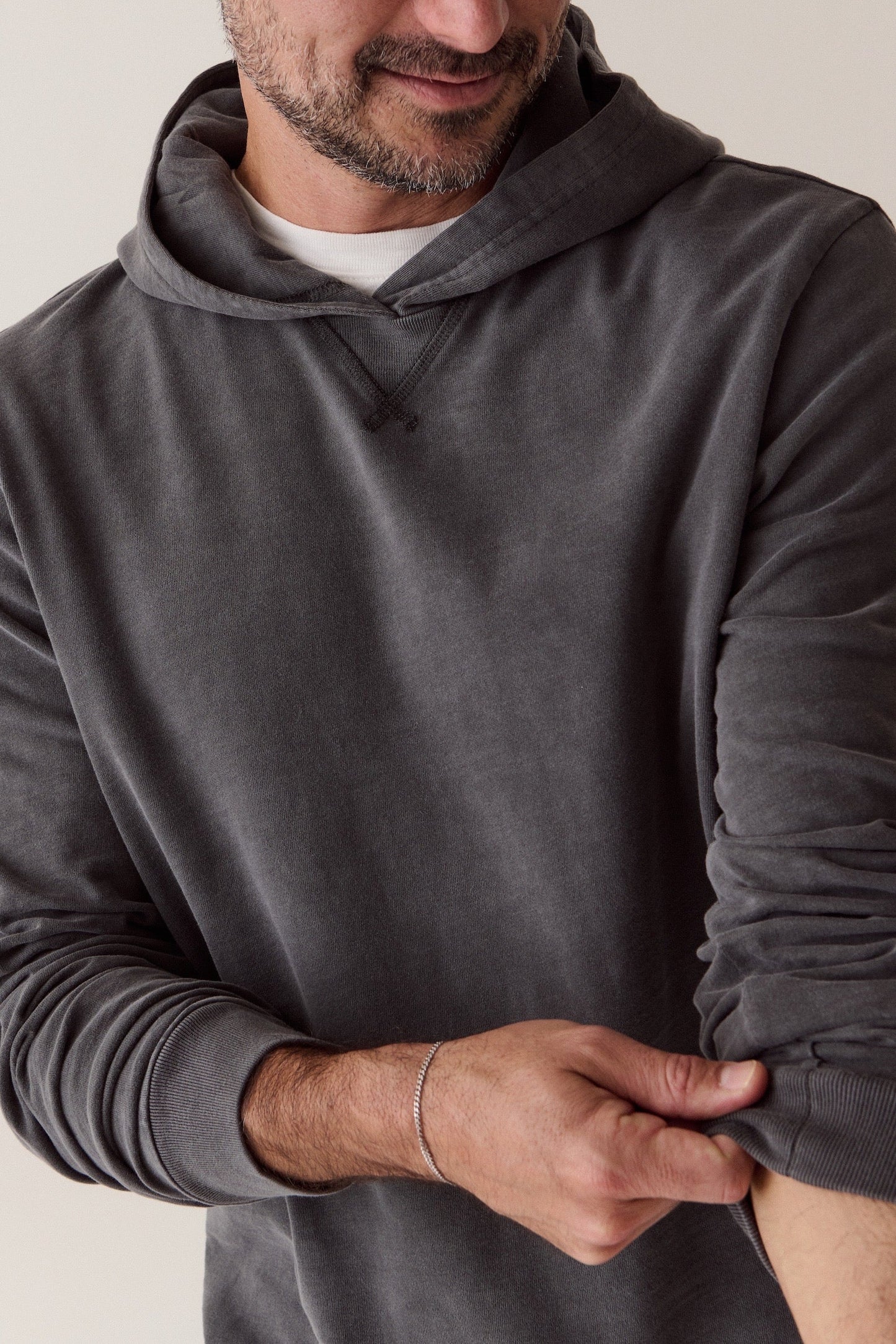 A man wearing a dark gray long-sleeve hoodie, shown from the chin down as he pulls up the sleeve of the garment. The background is a clean, neutral off-white studio wall.