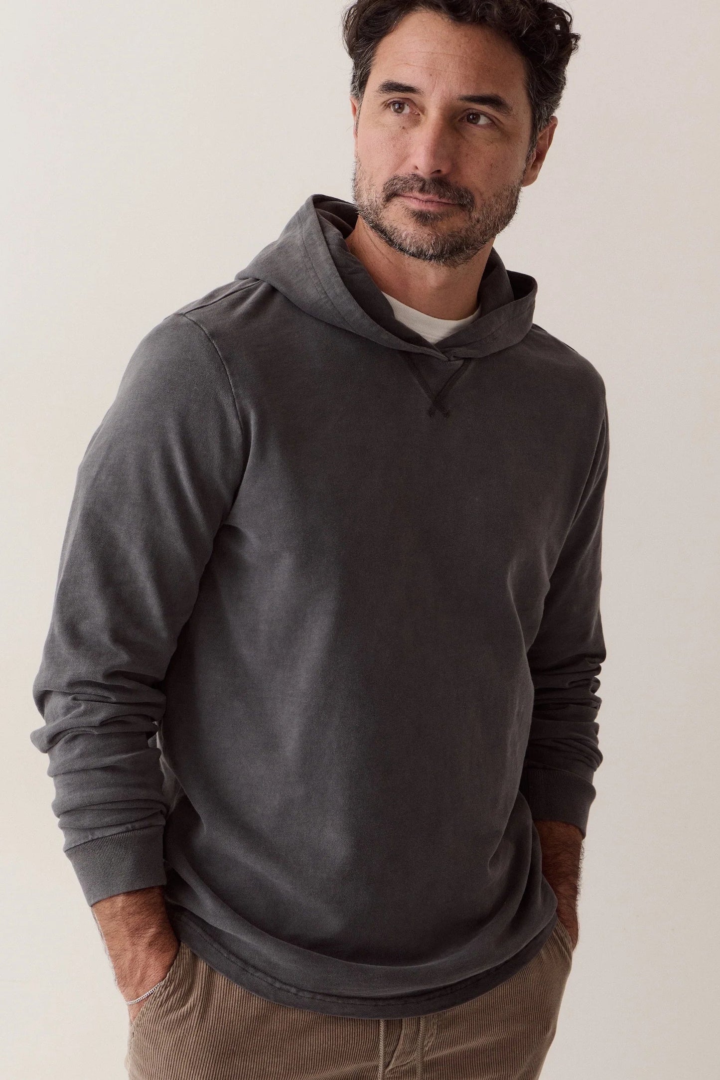 A man wearing a dark gray long-sleeve hooded sweatshirt over a white undershirt, paired with brown pants, standing against a plain light beige background.