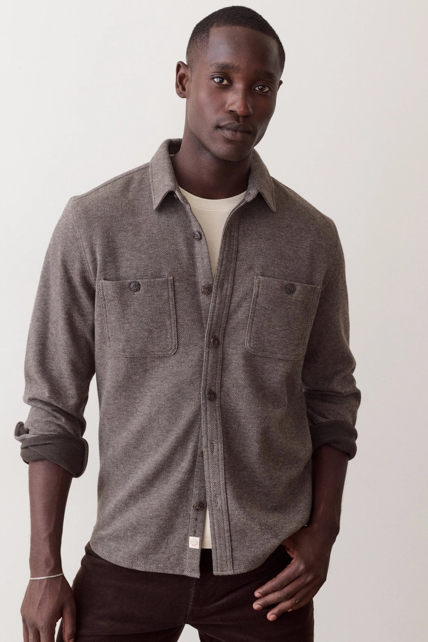 A man wearing a heather gray button-up shirt with two chest pockets over a cream undershirt and dark brown pants, standing against a plain off-white background.