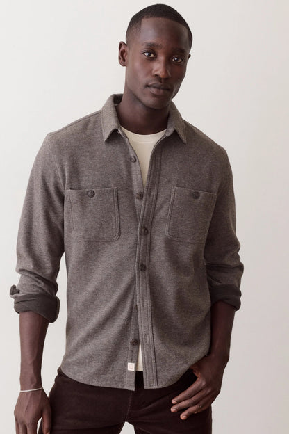A man wearing a heather gray button-up shirt with two chest pockets over a cream undershirt and dark brown pants, standing against a plain off-white background.