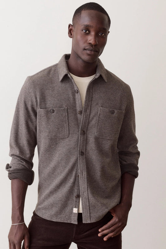 A man wearing a heather gray button-up shirt with two chest pockets over a cream undershirt and dark brown pants, standing against a plain off-white background.