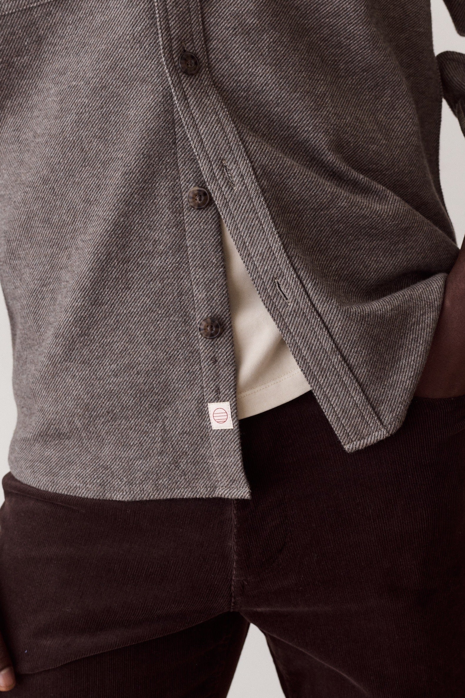 Close-up of a person wearing a textured gray button-front overshirt over a cream undershirt and dark brown corduroy pants, against a light neutral background.