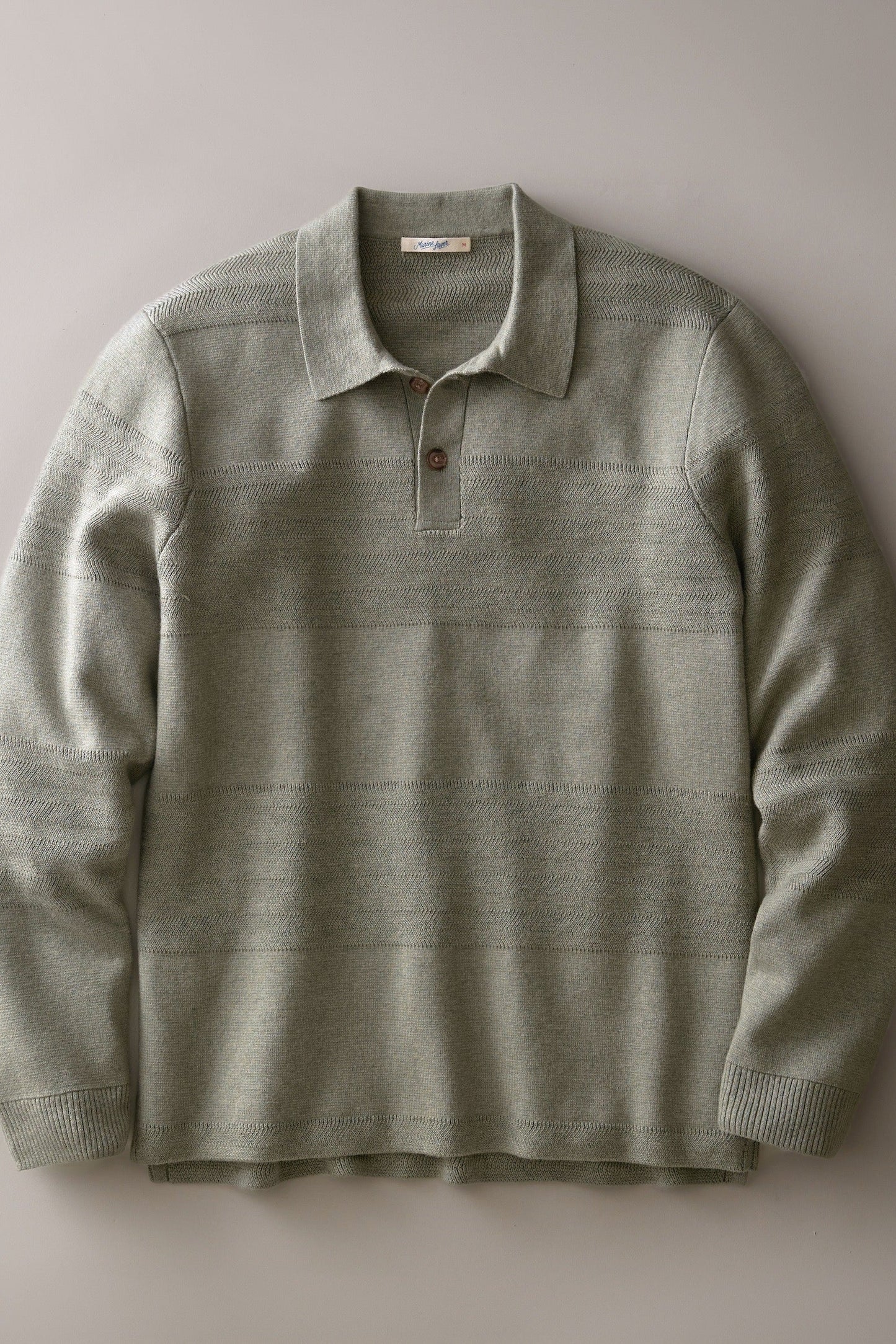 A long-sleeve knit polo shirt in a muted sage green color, featuring subtle horizontal textured stripes and a two-button placket, laid flat against a neutral gray background.