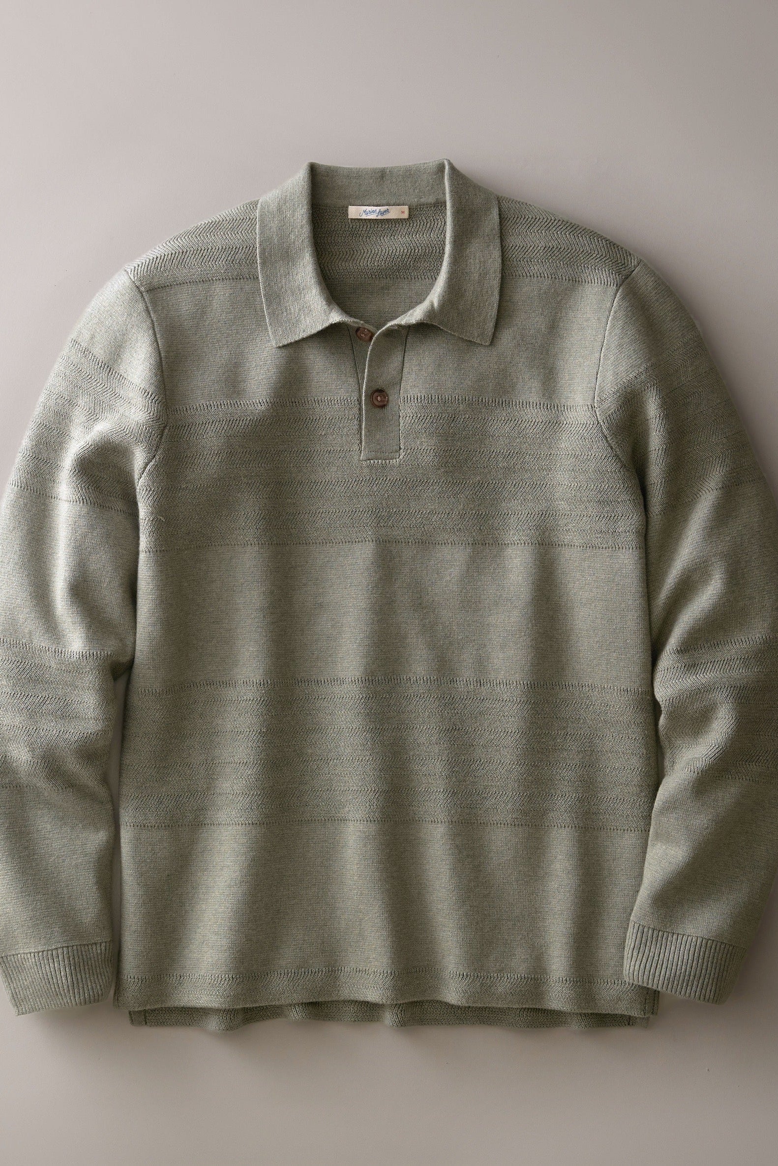 A long-sleeve knit polo shirt in a muted sage green color, featuring subtle horizontal textured stripes and a two-button placket, laid flat against a neutral gray background.
