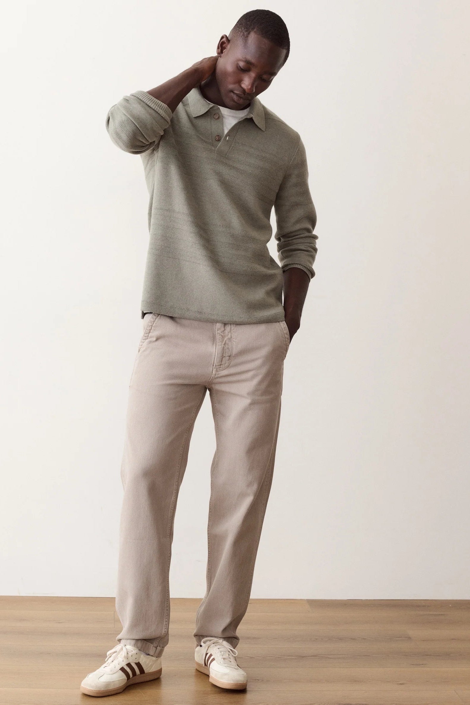A man wearing a light sage green knit polo sweater, beige trousers, and white sneakers with black stripes, standing against a plain off-white wall and light wood floor.
