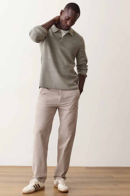A man wearing a light sage green knit polo sweater, beige trousers, and white sneakers with black stripes, standing against a plain off-white wall and light wood floor.