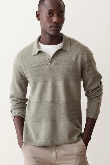A man wearing a light olive green long-sleeve knit polo sweater over a white t-shirt and beige trousers, standing against a plain off-white studio background.