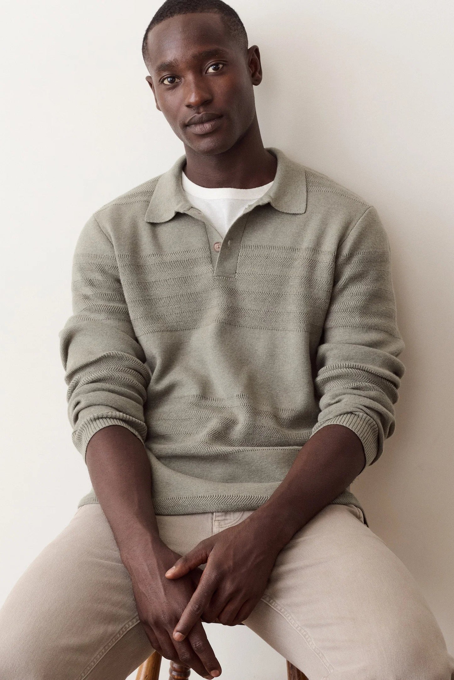 A man wearing a light sage green ribbed knit polo sweater over a white t-shirt and beige trousers, sitting against a plain off-white wall.