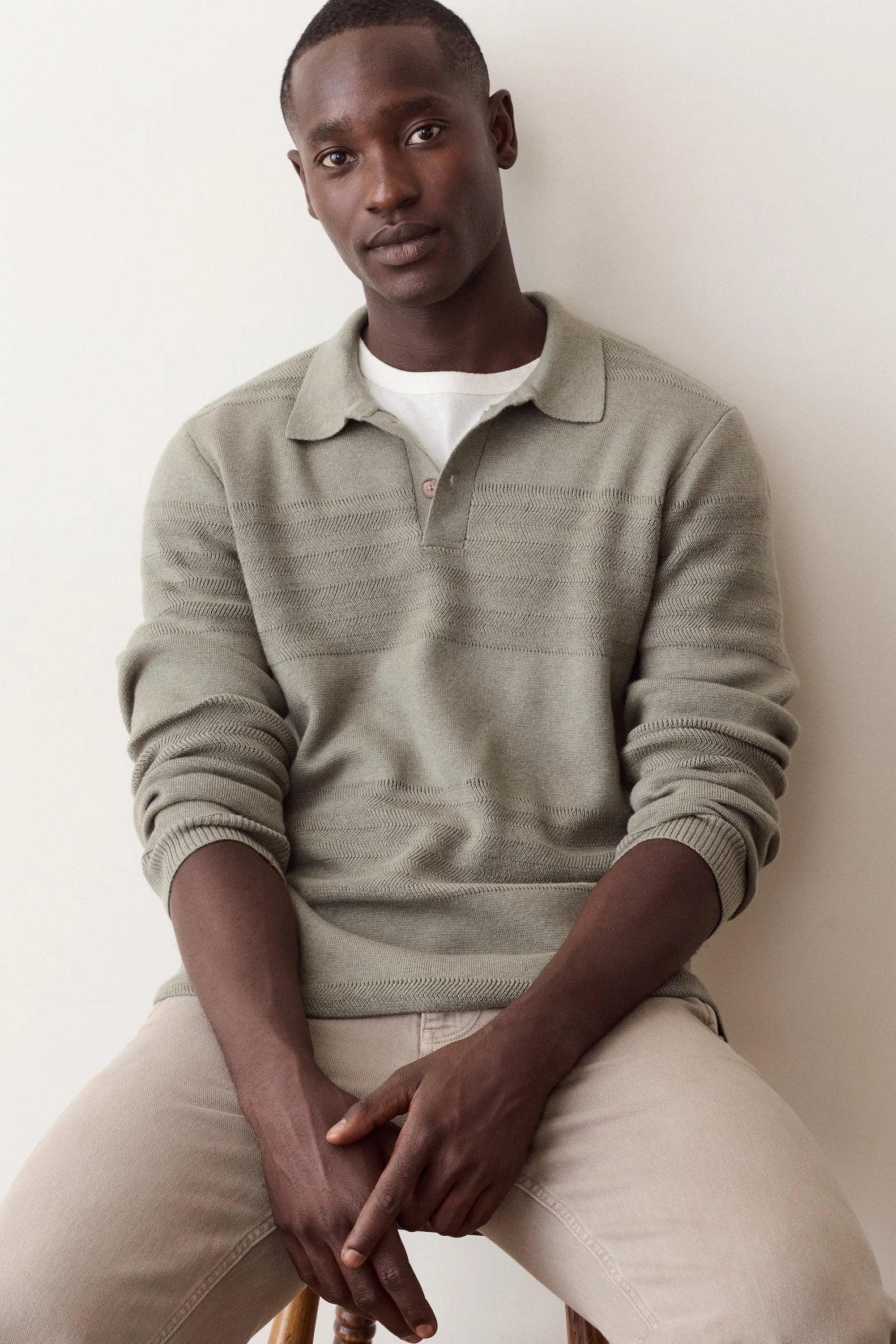 A man wearing a light sage green ribbed knit polo sweater over a white t-shirt and beige trousers, sitting against a plain off-white wall.