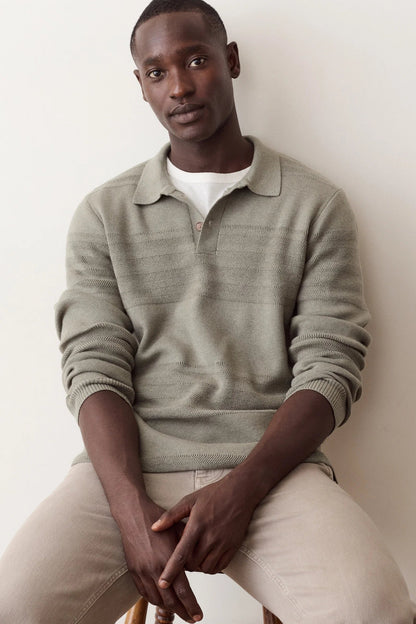 A man wearing a light sage green ribbed knit polo sweater over a white t-shirt and beige trousers, sitting against a plain off-white wall.