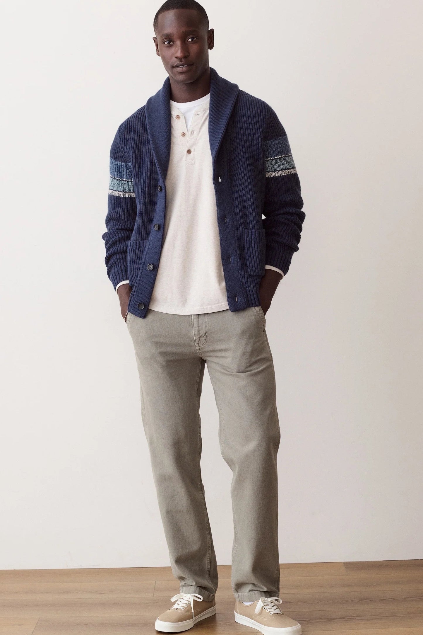A man modeling a navy blue ribbed knit cardigan with striped sleeve details, worn over a white henley t-shirt, paired with light gray trousers and beige sneakers, standing against a plain off-white wall.