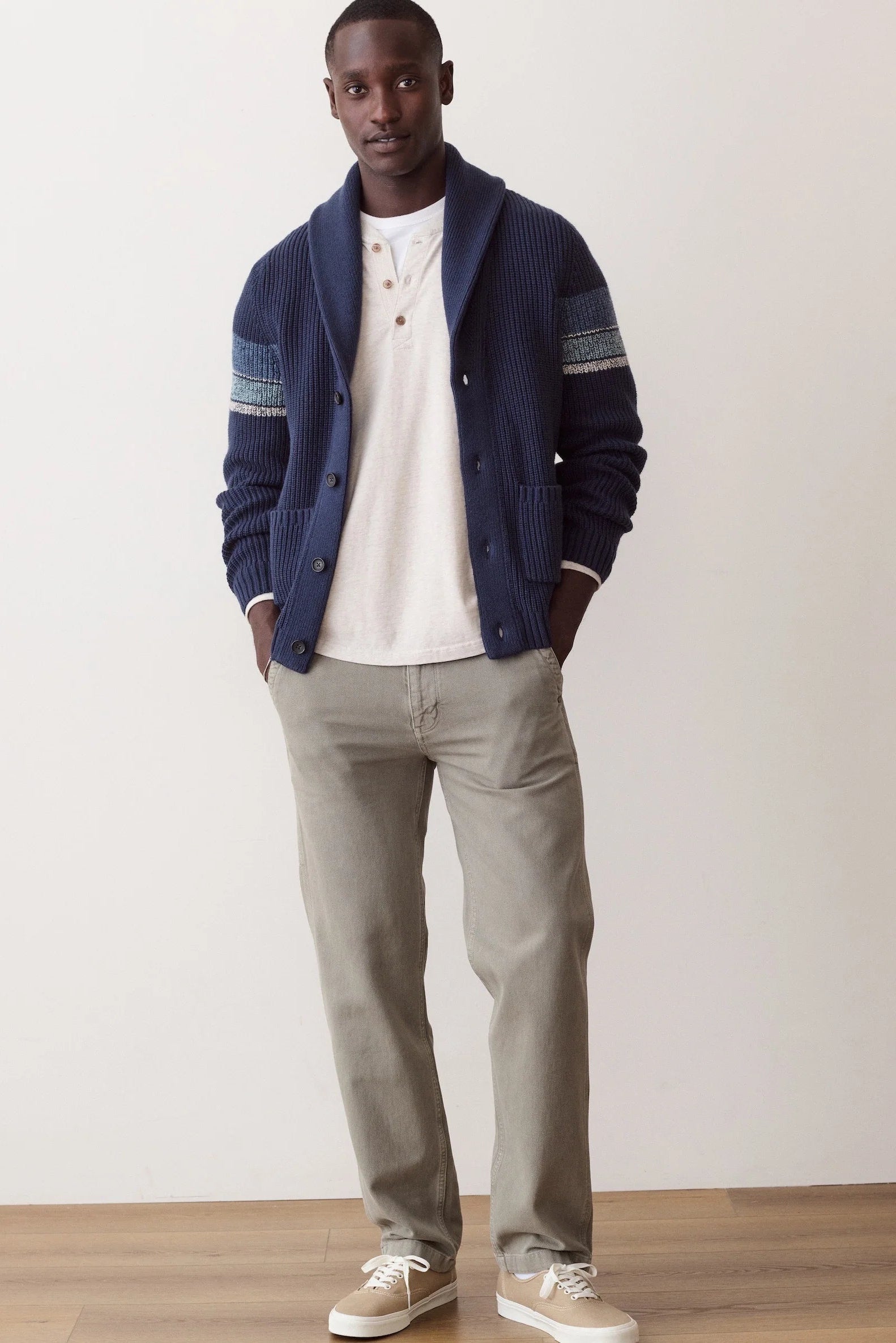A man modeling a navy blue ribbed knit cardigan with striped sleeve details, worn over a white henley t-shirt, paired with light gray trousers and beige sneakers, standing against a plain off-white wall.