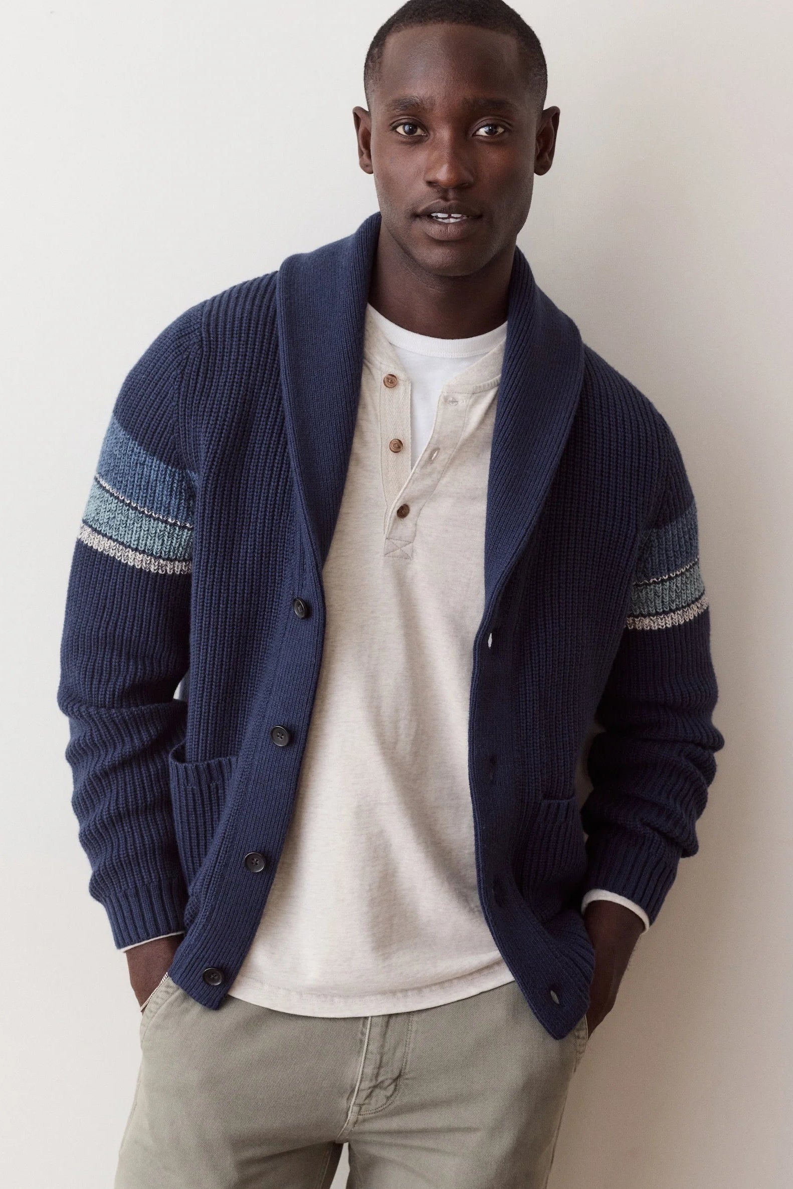 A man wearing a navy blue ribbed cardigan with textured blue and white sleeve stripes, layered over a cream henley shirt and khaki pants, standing against a plain off-white wall.