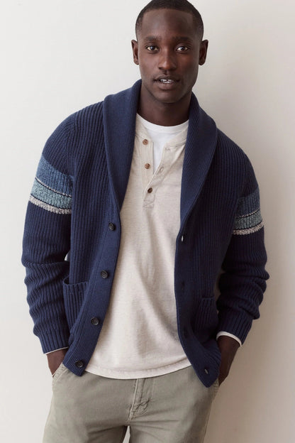 A man wearing a navy blue ribbed cardigan with textured blue and white sleeve stripes, layered over a cream henley shirt and khaki pants, standing against a plain off-white wall.