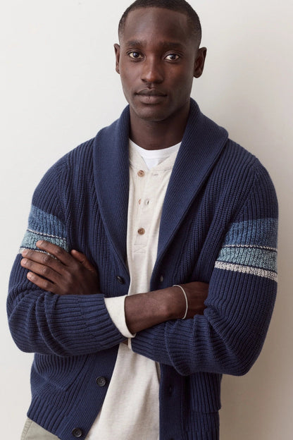 A man wearing a navy blue ribbed cardigan with textured blue and white knit sleeve stripes, layered over a cream henley shirt, standing against a plain off-white wall.