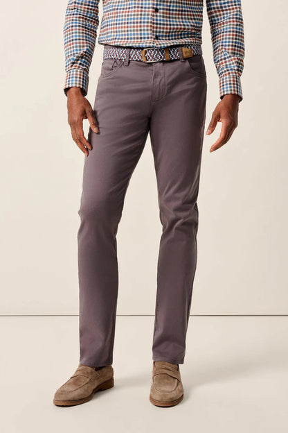 A man wearing slim-fit charcoal gray trousers, a multicolor checkered shirt, a patterned belt, and brown suede loafers, standing against a plain off-white studio background.