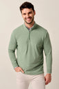 A smiling man wearing a sage green long-sleeve quarter-zip pullover sweater over a patterned collared shirt and white pants, standing against a plain light beige studio background.