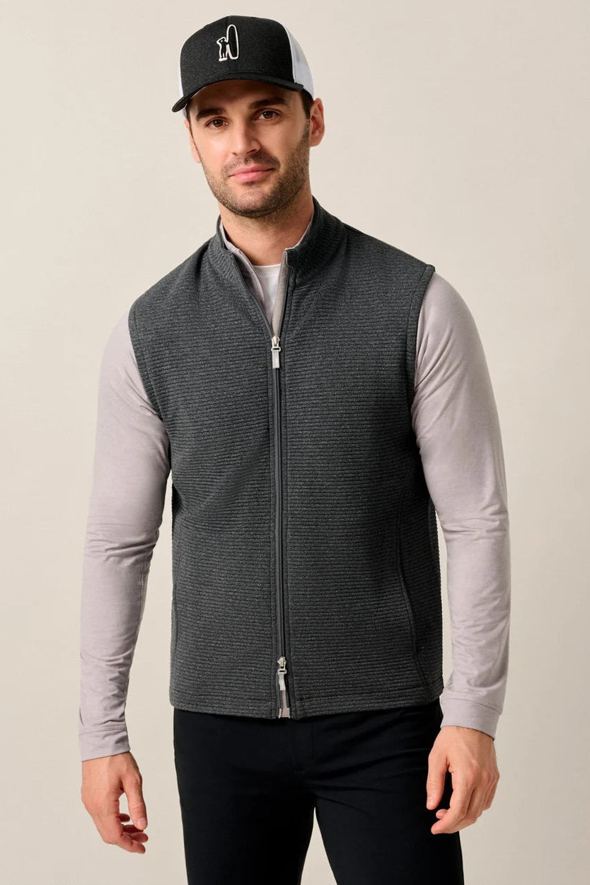 Man wearing a gray vest over a light gray long-sleeve shirt with a black cap on a beige background