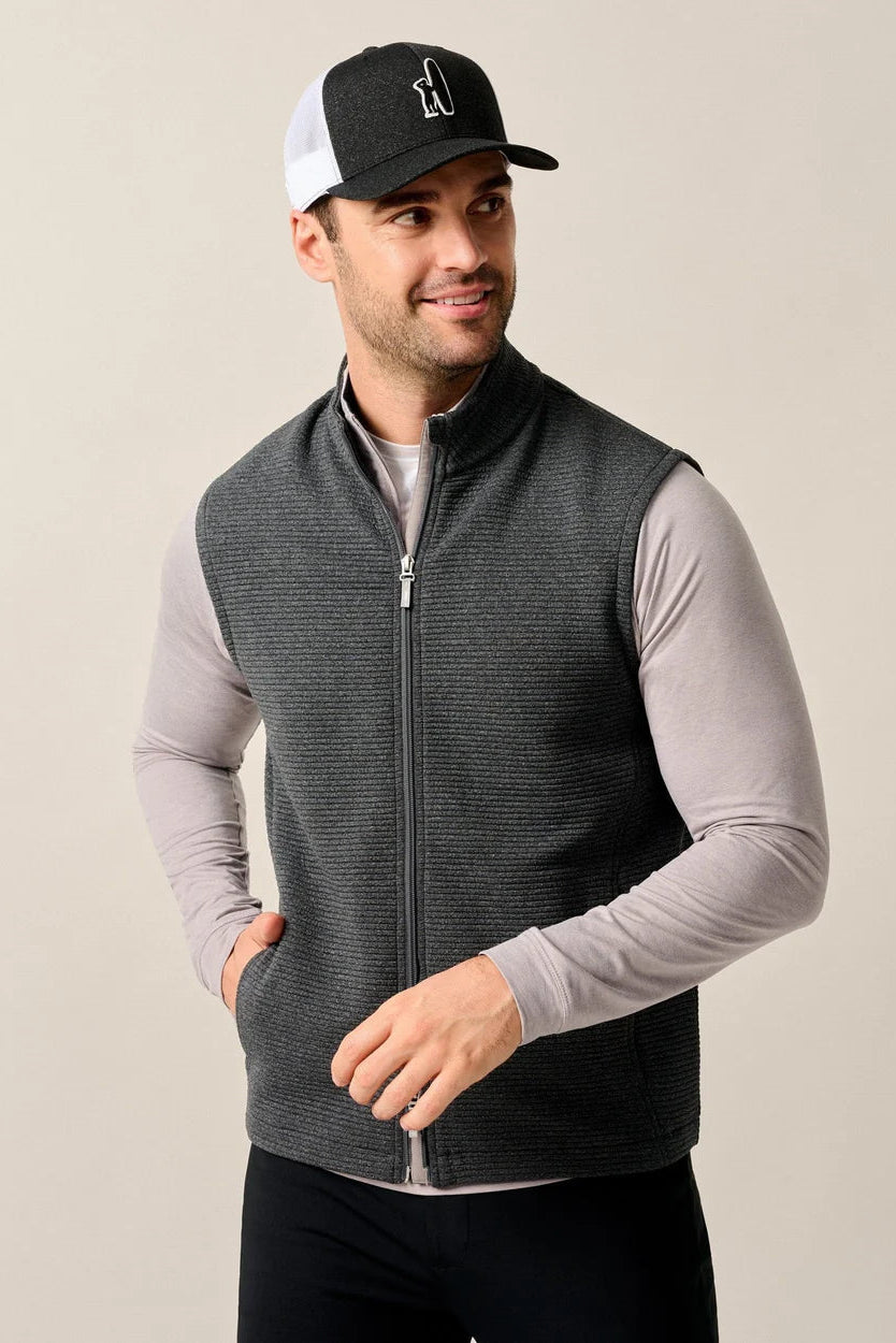 A man wearing a dark gray zip-up knit vest over a light gray long-sleeve shirt, paired with a black and white trucker hat, against a plain beige studio background.