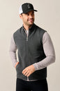 A man wearing a dark gray zip-up knit vest over a light gray long-sleeve shirt, paired with a black and white trucker hat, against a plain beige studio background.