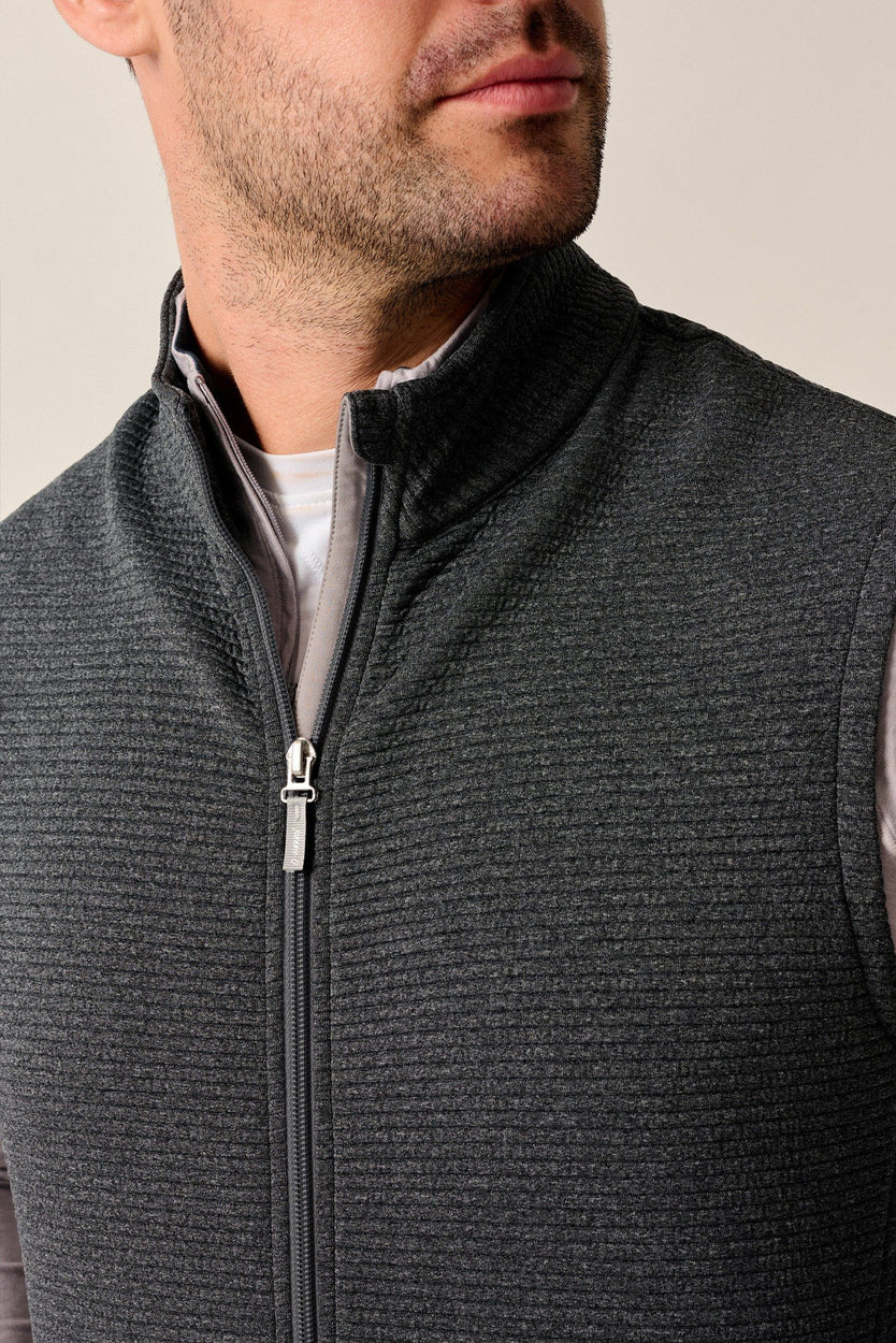 A close-up of a man wearing a dark gray ribbed zip-up vest over a light gray shirt, cropped from the chin down against a solid light gray background.