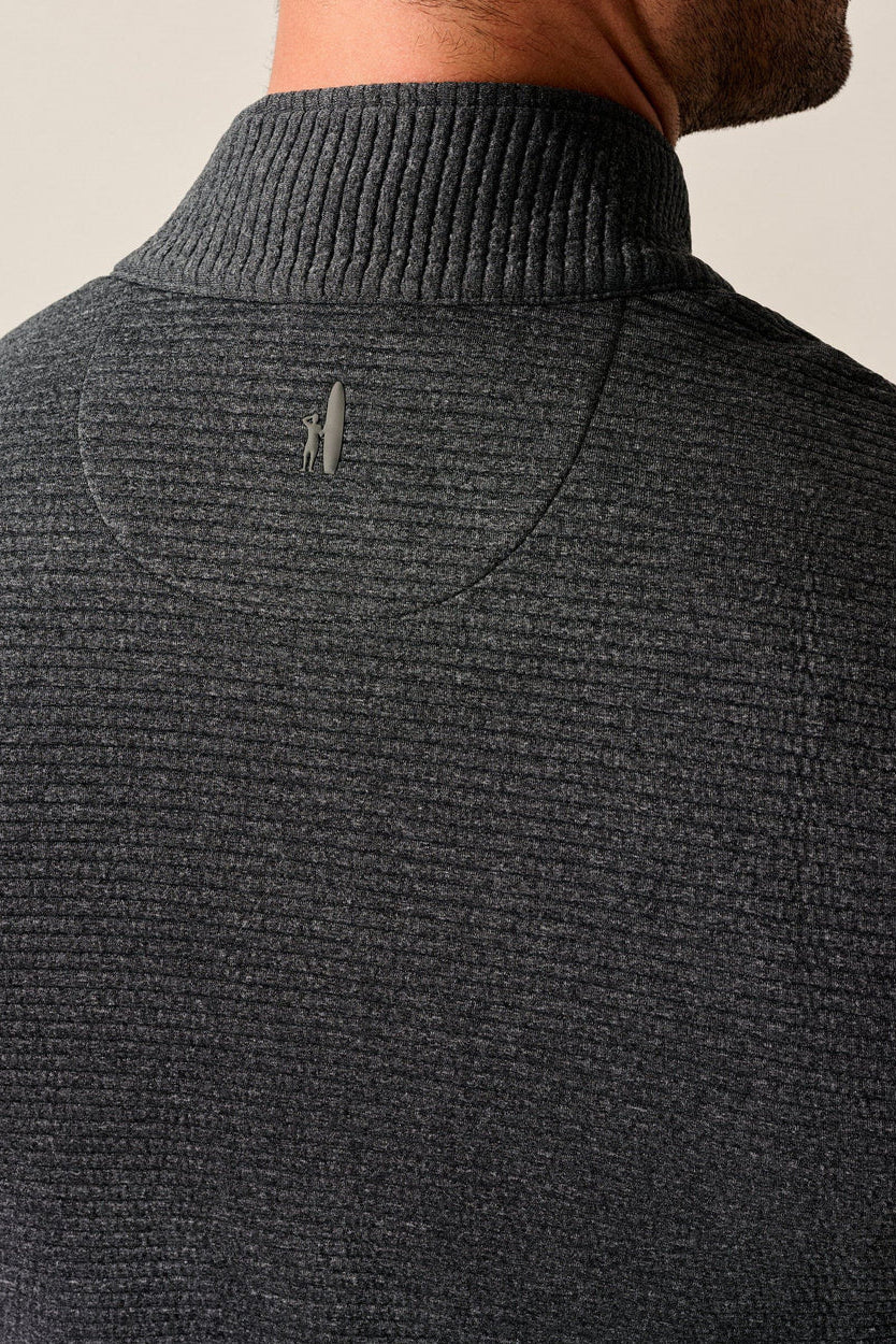 Close-up back view of a man wearing a dark charcoal gray ribbed knit mock neck sweater, showing a small metallic logo on the upper back.
