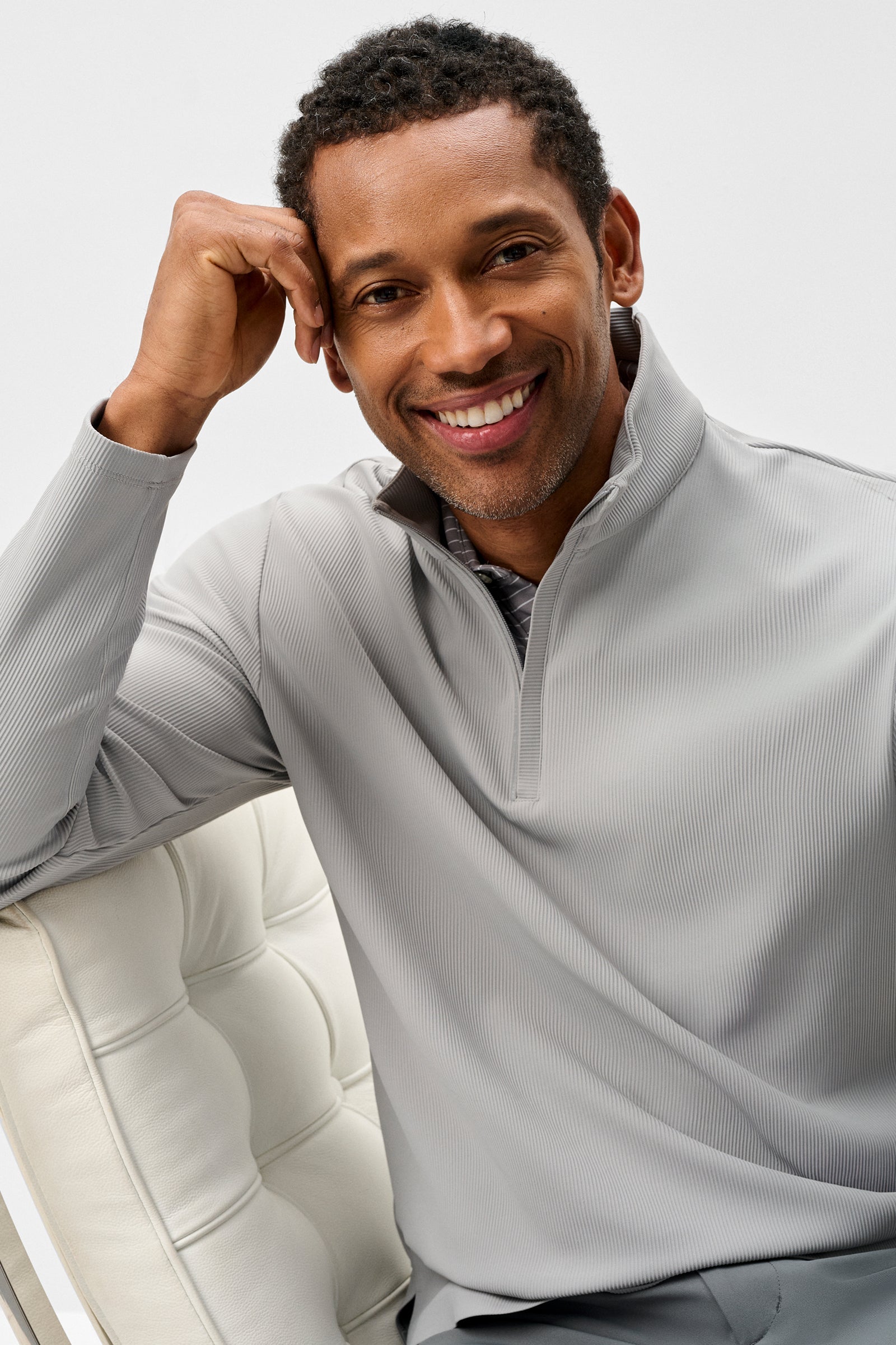 Man wearing a light gray pullover sitting on a white chair against a plain background