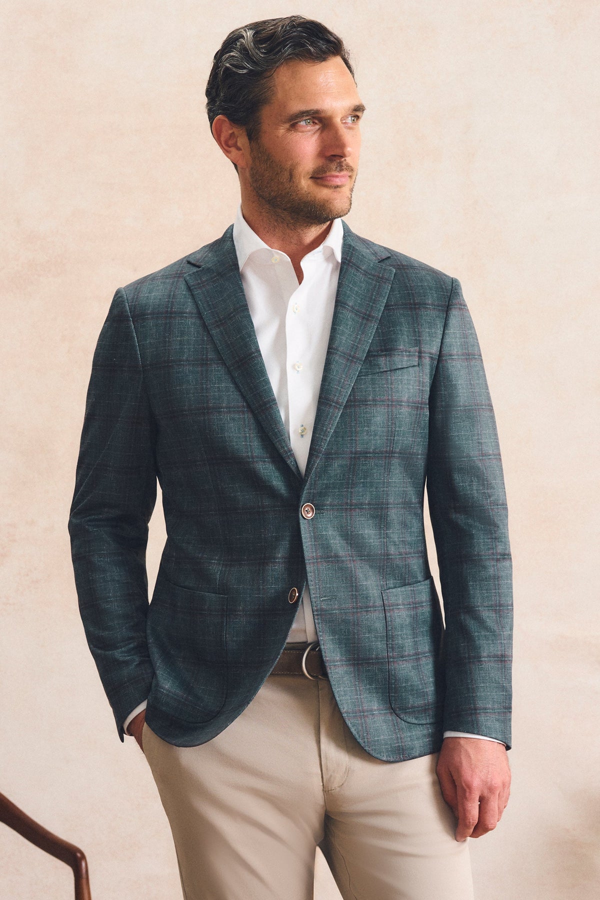 A man wearing a dark teal and gray plaid blazer over a white dress shirt and beige trousers, standing against a textured beige wall.