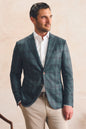 A man wearing a dark teal and gray plaid blazer over a white dress shirt and beige trousers, standing against a textured beige wall.
