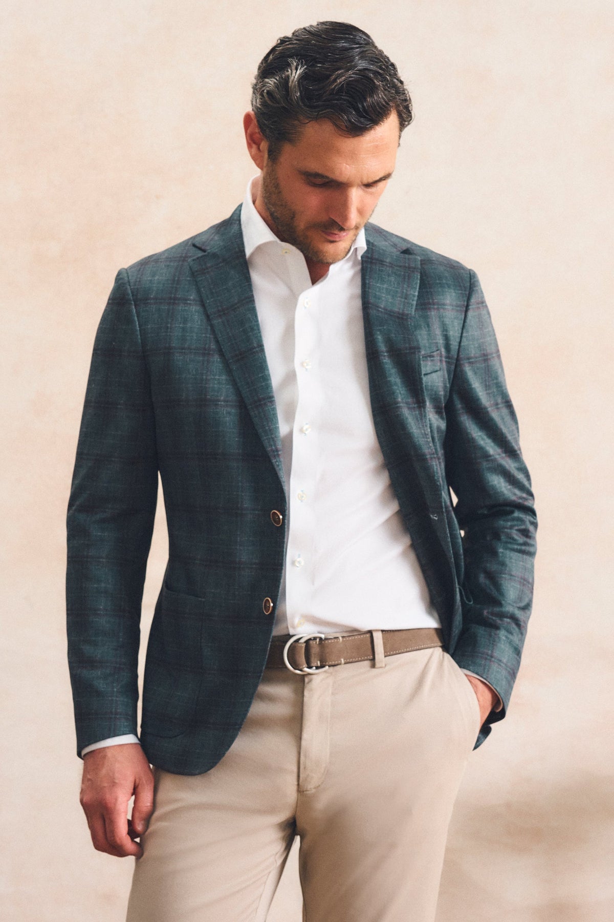 A man wearing a dark teal-green plaid blazer over a white dress shirt, paired with beige trousers and a brown belt, standing against a textured beige wall.