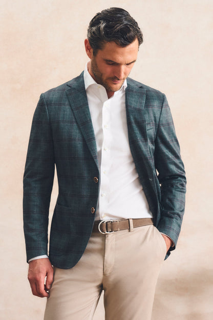 A man wearing a dark teal-green plaid blazer over a white dress shirt, paired with beige trousers and a brown belt, standing against a textured beige wall.