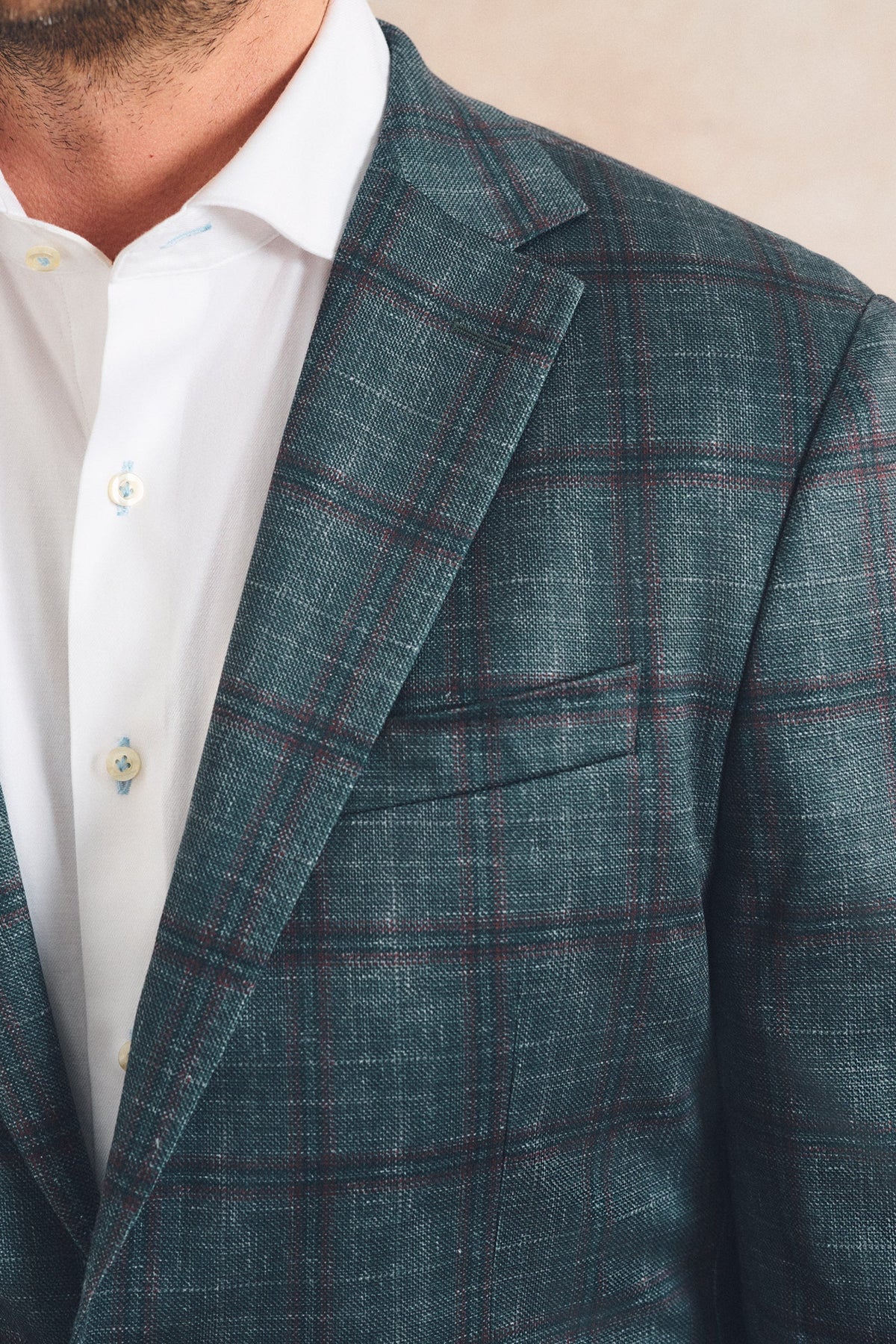 A close-up of a man wearing a teal blue-green plaid blazer over a white button-down shirt. The blazer features a subtle windowpane check pattern in muted red and brown lines.
