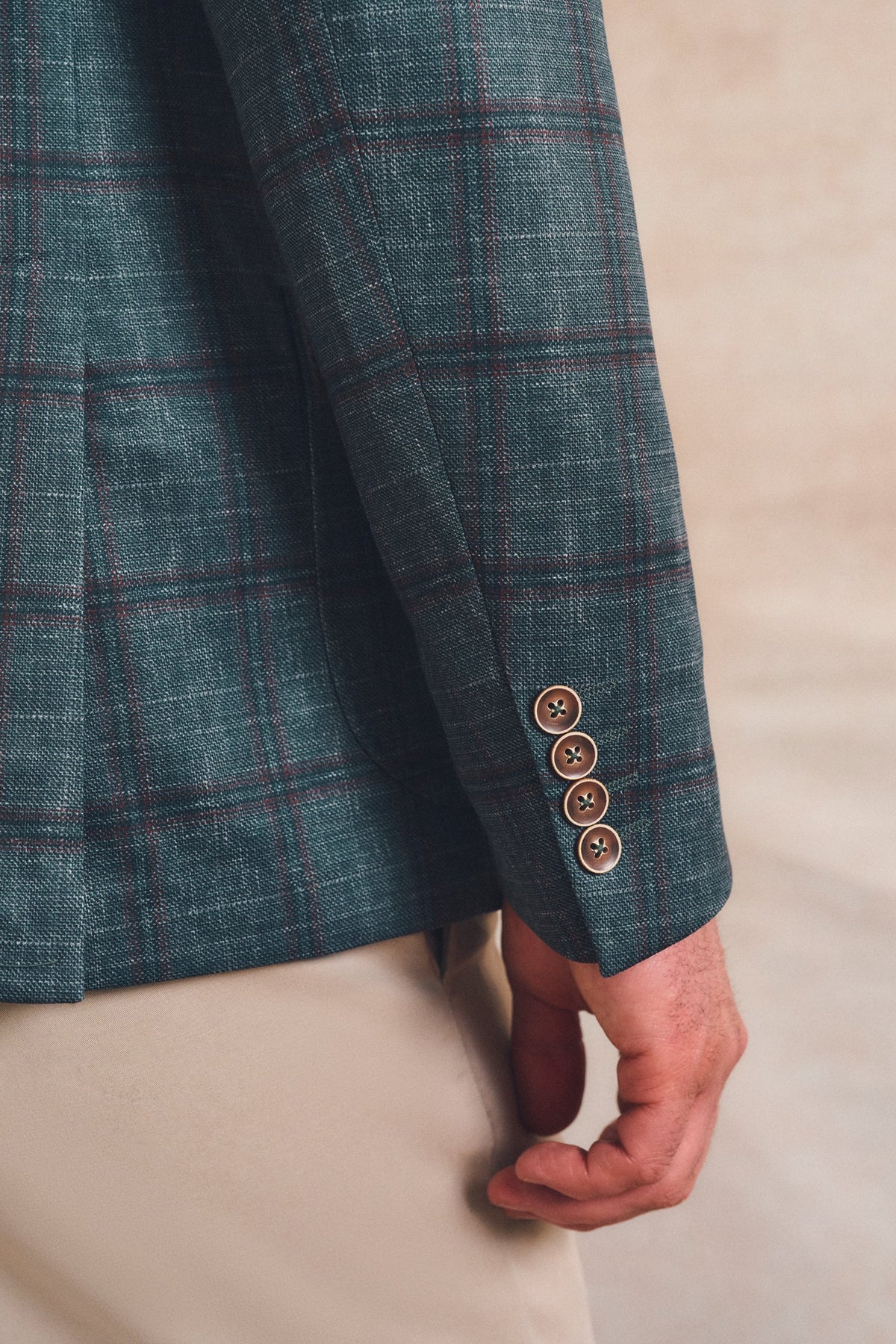 Close-up of a person wearing a teal plaid blazer with brown buttons on the cuff, paired with beige trousers, against a neutral beige background.