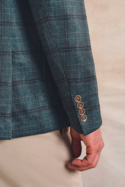Close-up of a person wearing a teal plaid blazer with brown buttons on the cuff, paired with beige trousers, against a neutral beige background.
