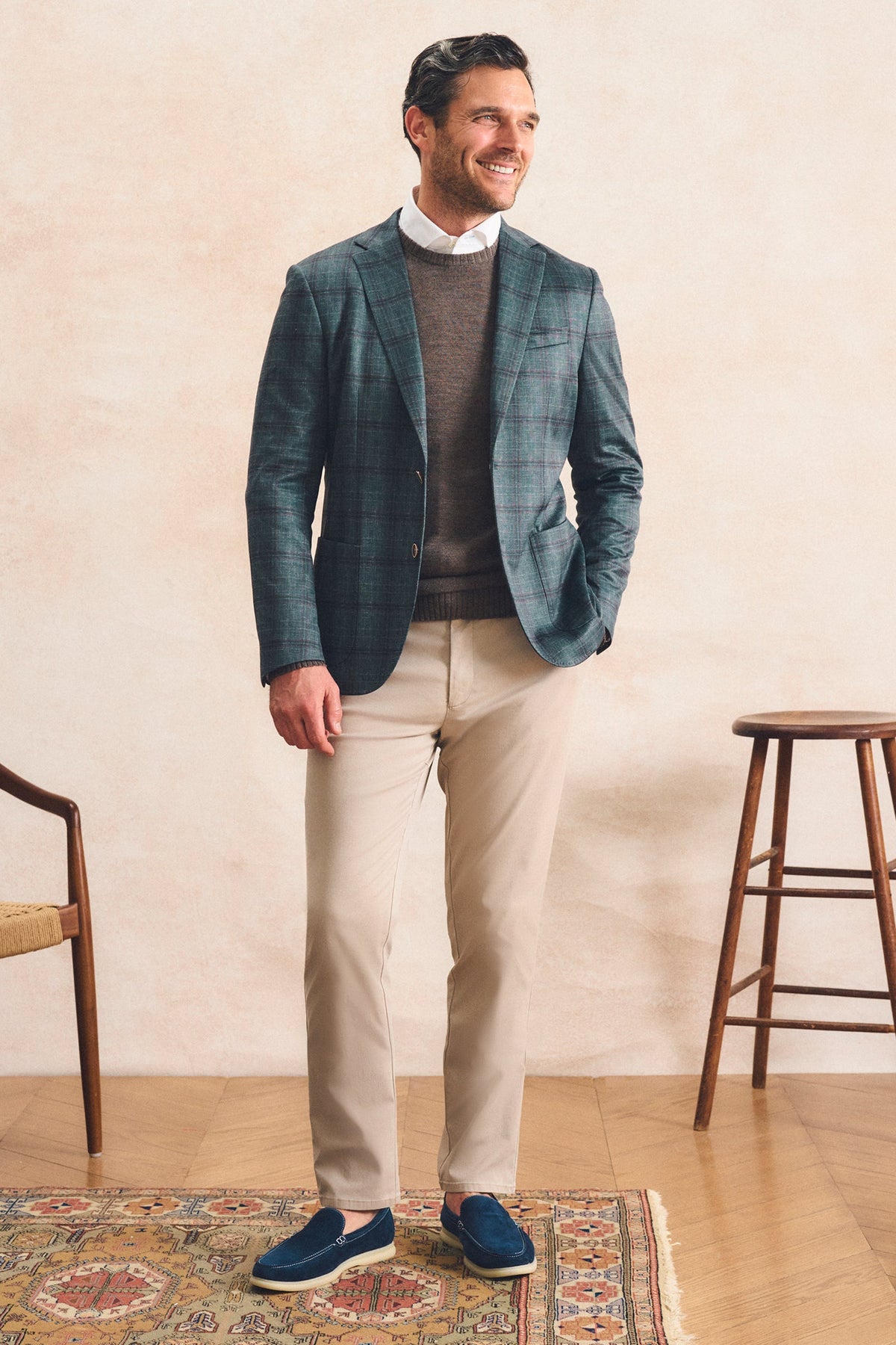 A man stands in a styled indoor setting wearing a teal and navy plaid blazer over a brown sweater, a white collared shirt, beige trousers, and blue slip-on shoes. A patterned rug, wooden stool, and chair are visible against a textured beige wall.