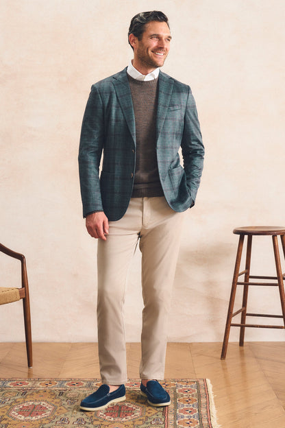 A man stands in a styled indoor setting wearing a teal and navy plaid blazer over a brown sweater, a white collared shirt, beige trousers, and blue slip-on shoes. A patterned rug, wooden stool, and chair are visible against a textured beige wall.