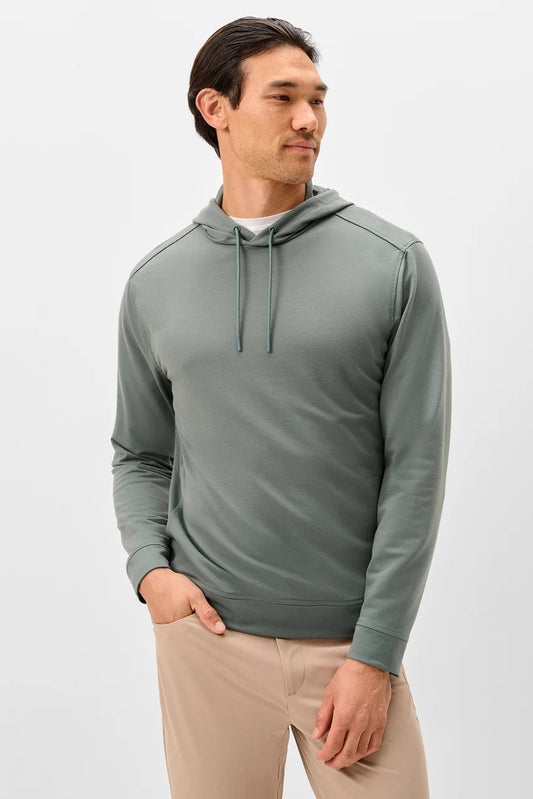 A man wearing a sage green long-sleeve athletic hoodie with drawstrings, paired with beige pants, standing against a light gray studio background.