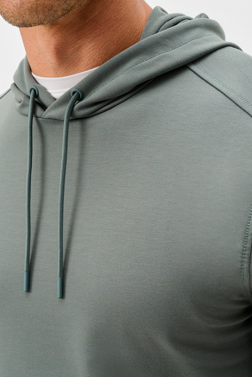Close-up of a person wearing a muted sage green hooded sweatshirt with matching drawstrings over a white undershirt, against a light gray background.